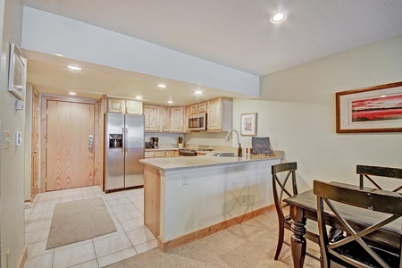 A modern kitchen with wooden cabinets, stainless steel appliances, a countertop bar, and recessed lighting. A dining area with a wooden table and chairs is situated to the right.