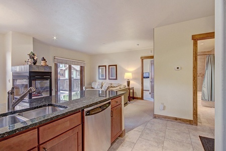 A modern, open-plan kitchen and living room area with granite countertops, wood cabinets, a stainless steel dishwasher, a fireplace, and a partially visible bedroom entrance on the right.