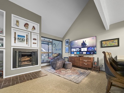 A living room with a TV mounted on the wall, entertainment center, fireplace, and various wall art. There's a rug, chair, and seating. Windows let in natural light and showcase an outdoor view.