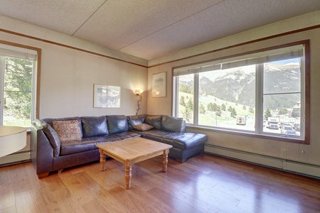 A living room with a brown sectional couch, wooden coffee table, hardwood floor, and large window offering a mountain view.