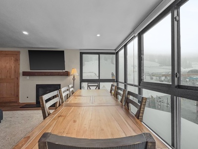 A dining area with a wooden table and chairs is adjacent to large windows offering a snowy mountain view. A fireplace and a wall-mounted TV are on the left side.