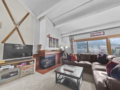 A living room with a brown sectional couch, a fireplace, a TV on a wooden stand, and a large window showcasing a mountain view. Skis are mounted on the wall above the fireplace.