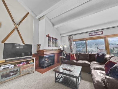 A living room with a brown sectional couch, a fireplace, a TV on a wooden stand, and a large window showcasing a mountain view. Skis are mounted on the wall above the fireplace.