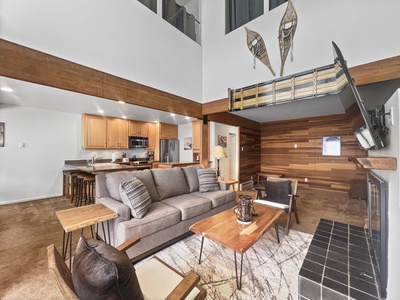 A living room with a gray couch, wooden tables, and chairs. The open space extends to a kitchen. A staircase leads to an upper level with wooden snowshoes mounted on the wall.