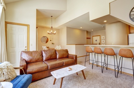 A living room with a brown leather sofa, a small coffee table, and a blue armchair. A kitchen with a breakfast bar and four bar stools. A dining area with a table and chairs is visible in the background.