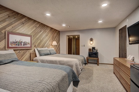 A bedroom with two double beds, wood-paneled wall, wall-mounted TV, dresser, and a small bench holding a microwave and coffee maker. The room is lit by recessed lighting.