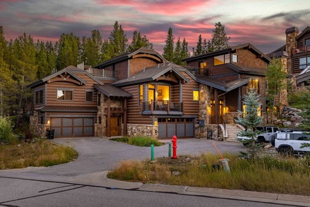 Large, multi-story wooden house with lush greenery, a driveway, and multiple garages under a vibrant sky.