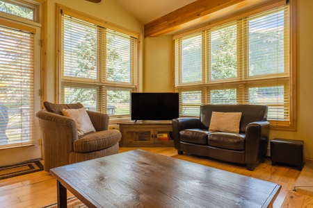 The living room offers a TV and multiple seating options. Large windows allow ample natural light to come in.