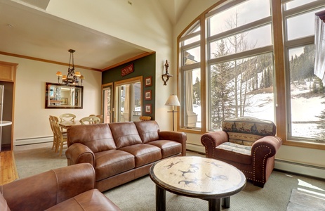 A cozy living room features brown leather seating, a round coffee table, large windows with a snowy mountain view, and a dining area with wooden chairs in the background.