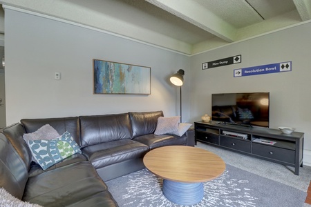 A modern living room with a black leather sectional sofa, a round wooden coffee table, a flat-screen TV on a media console, and abstract wall art. Decorative signs and a floor lamp are present.