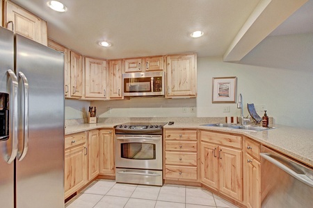 Modern kitchen with light wood cabinetry, stainless steel appliances, granite countertops, and recessed lighting. Includes a refrigerator, oven, microwave, and an undermount sink with faucet.