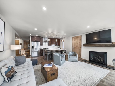 A modern living area with a light gray sofa, wooden coffee table, two gray armchairs, a flat-screen TV over a fireplace, and an open kitchen with dark cabinets and stainless steel appliances.