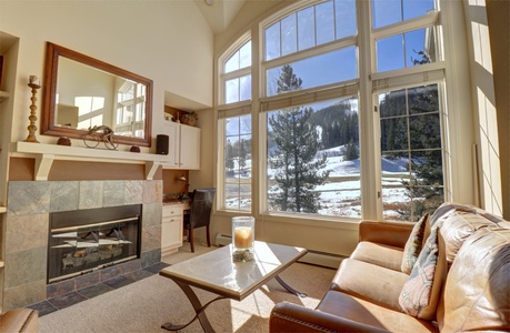 Spacious living room with large windows offering a view of the snowy East Village ski slopes. Features a fireplace, a cozy sofa, and coffee table. Natural light fills the room.