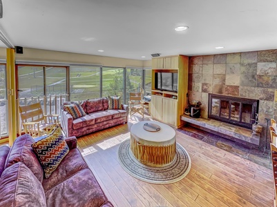 Modern living room with large windows, two purple sofas, a round coffee table, a fireplace with stone surround, and a wall-mounted TV. Wood flooring and a few decorative pillows accent the space.