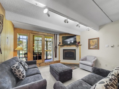 A cozy living room with gray sofas, a fireplace with a mounted TV, and a wooden wall. Large windows and doors allow in natural light, and a framed painting hangs on the wall.