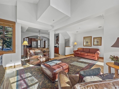 A well-lit, spacious living room with leather furniture, a large area rug, a wooden coffee table, and adjacent open kitchen area. Framed pictures hang on the walls, and there is a staircase in the background.