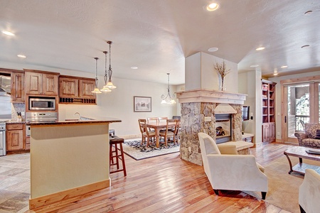 A spacious open-plan kitchen and living area with wooden floors, a stone fireplace, a dining table, modern kitchen appliances, and large windows letting in natural light.