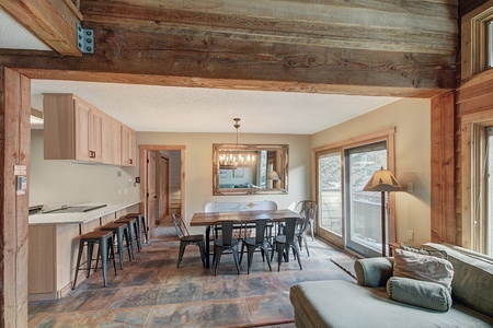 Open-concept kitchen and dining area with a wooden table, metal chairs, a large mirror, and a windowed door leading outside. The space features rustic wooden beams and a neutral color palette.