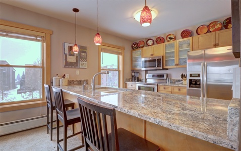 A modern kitchen with wooden cabinets, granite countertops, bar seating, stainless steel appliances, and decorative plates above cabinets. Large windows allow natural light to enter.