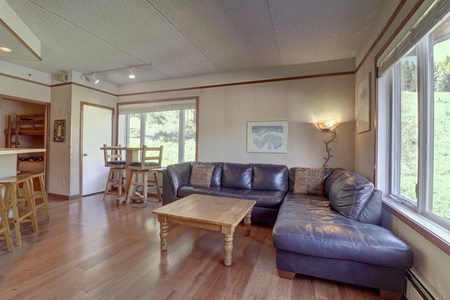 A cozy living room with a dark leather sectional sofa, a wooden coffee table, stools by a kitchen counter, a wall-mounted light fixture, and large windows offering an outdoor view.