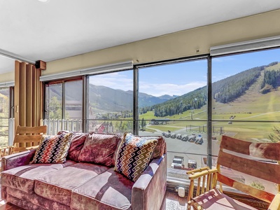 A living room with a purple couch, wooden chairs, and large windows showcasing a scenic mountain view on a sunny day.