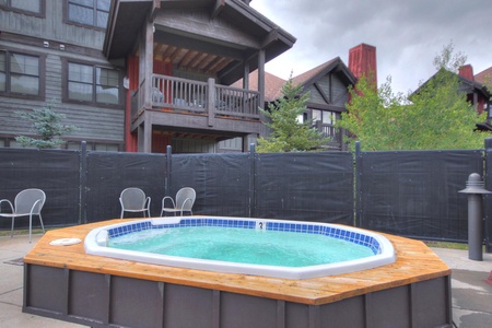 Outdoor hot tub surrounded by wooden decking with several chairs nearby, set against the backdrop of multi-story rustic buildings with balconies and overcast skies.