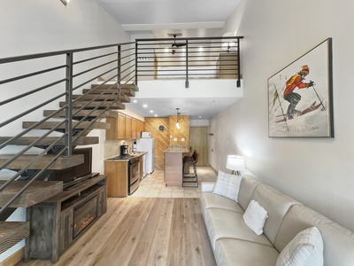 A modern loft apartment with a staircase, kitchenette, and living area. There is a sofa, fireplace, and large wall art featuring a skier. The upstairs area has a metal railing.