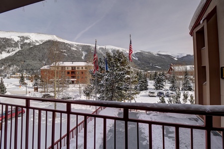 A snow-covered mountain landscape with buildings and trees, viewed from a balcony with three flags on poles. The scene includes parked cars and a partially cloudy sky.