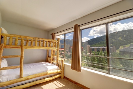 A room with a wooden bunk bed next to large windows offering a view of mountains and buildings under a partly cloudy sky. The room is well-lit with natural light.