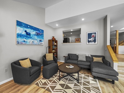 A modern living room with a gray sectional sofa and two gray armchairs around a round wooden coffee table. The room features light wood flooring, framed artwork, and built-in bookshelves.