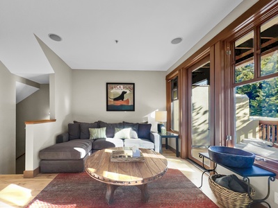 A cozy living room with a gray sectional sofa, round wooden coffee table, and a dog-themed wall art. Large windows let in ample natural light and provide a view of greenery outside.