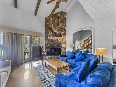 Spacious living room with high ceilings, stone fireplace, blue sofas, wooden coffee table, large window with a view of trees, and an adjacent staircase.