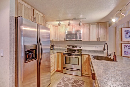 Modern kitchen with stainless steel appliances including a double-door refrigerator, oven, and microwave. Light wood cabinets, a sink with a faucet, and a gray countertop. Track lighting overhead.