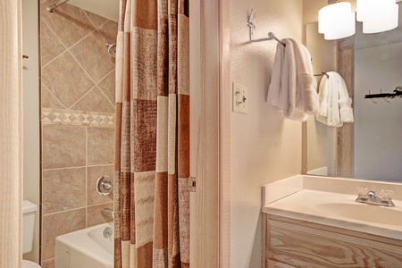 Bathroom with a toilet, a bathtub with tiled walls, a shower curtain, a vanity with a sink, and a mirror above. Two white towels hang on a wall-mounted towel rack.