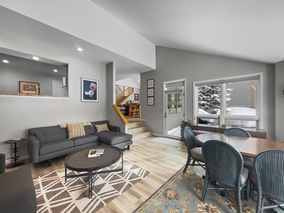 A modern living and dining area features gray sofas, a round coffee table, a dining table with blue chairs, and decor on walls. Large windows reveal a snowy outdoor scene. Stairs lead to a second level.