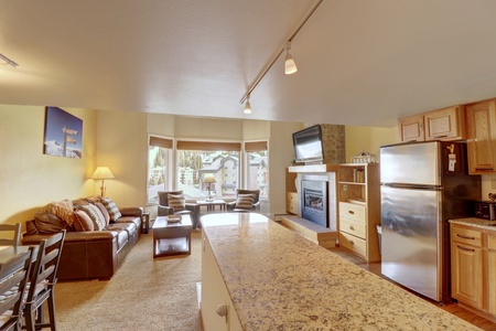 A modern apartment interior featuring a living room with a leather sofa, dining area, kitchen with stainless steel appliances, and a large window. A fireplace and mounted TV are visible.