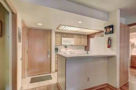 A small kitchen area with light wooden cabinets, white appliances including a microwave and refrigerator, a countertop bar, and an adjacent wooden front door. A fire extinguisher is mounted on the wall.