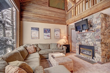 Cozy living room with a large sectional sofa, stone fireplace, flat-screen TV, and wall art. A window reveals a snowy outdoor scene, and a wooden loft overlooks the space.