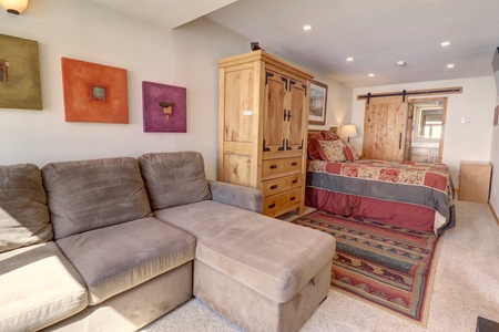 A cozy bedroom features a sofa, a wooden armoire, a bed with patterned bedding, and a colorful rug. Framed artwork decorates the walls, and there is a sliding wooden door in the background.