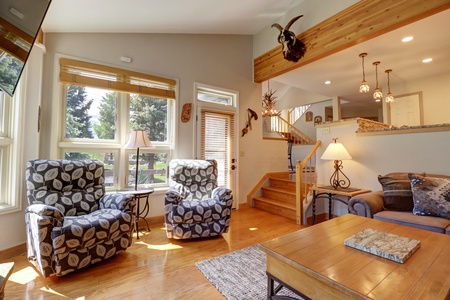 A cozy living room with two patterned armchairs, a wooden coffee table, and a couch. A staircase leads to an upper level with hanging pendant lights and wall-mounted decor. Large windows let in natural light.