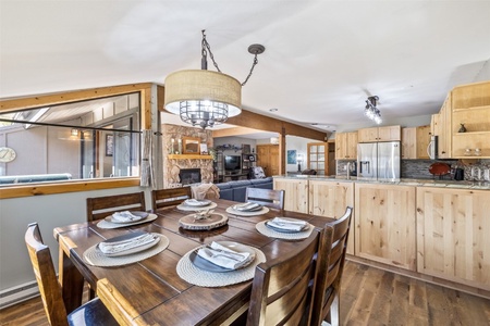 A cozy dining area with a wooden table set for six, adjacent to an open kitchen with light wood cabinets and stainless steel appliances. A stone fireplace is visible in the living area in the background.