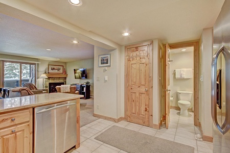 A basement with a kitchen area on the left, living space in the background, a wooden door leading to a bathroom on the right, and a stainless steel refrigerator on the far right.