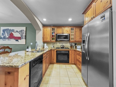 A modern kitchen with wooden cabinets, stainless steel refrigerator, granite countertops, built-in microwave, and a dishwasher. A painting of a red barn and snowy landscape hangs on the green wall.