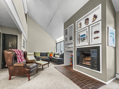 A cozy living room features a leather armchair, an L-shaped sofa, a fireplace with decorated shelves above it, and a brick hearth. Adjacent walls are adorned with artwork and photographs.