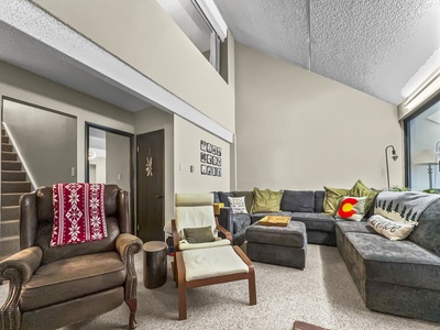 A cozy living room features a sectional sofa with green and yellow pillows, several armchairs, a wall-mounted grid of small frames, and a staircase leading to the upper floor.