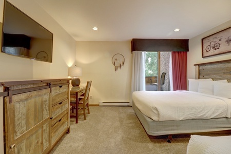 A hotel room with a bed, TV, wooden dresser, desk with a chair, and a large window with curtains. The room is carpeted and features a dreamcatcher and bicycle-themed wall art.