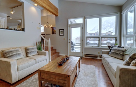 A cozy living room with two beige sofas, a wooden coffee table, and a view of snow-capped mountains through large windows. The room has white walls and wooden floors.