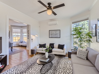 A modern living room with a white sofa, gray couch, black coffee table, ceiling fan, potted plants, and patterned rug. Adjacent room visible through a wide doorway.