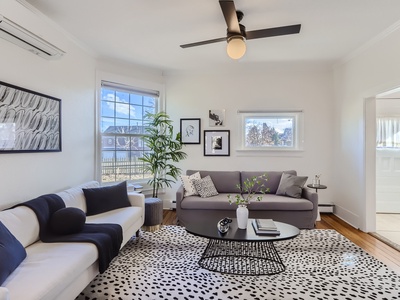 A modern living room with a white couch, grey sofa, black and white rug, black coffee table, potted plant, wall art, and a ceiling fan. A large window and a small window allow natural light in.