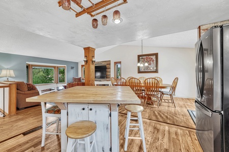 Open-concept kitchen and dining area with a wooden island and stools in the foreground, dining table with chairs in the background, and a sectional sofa with a TV in the living area on the left.